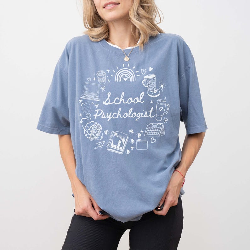 May include: A blue short-sleeved t-shirt with the words "School Psychologist" in white script, surrounded by a circular design of a laptop, coffee cup, rainbow, and other graphics. The shirt is made of a soft material.