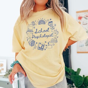 May include: A light yellow t-shirt with a blue graphic that says "School Psychologist." The graphic includes a laptop, coffee cup, rainbow, and other school-related icons. The shirt is a comfortable, oversized fit.