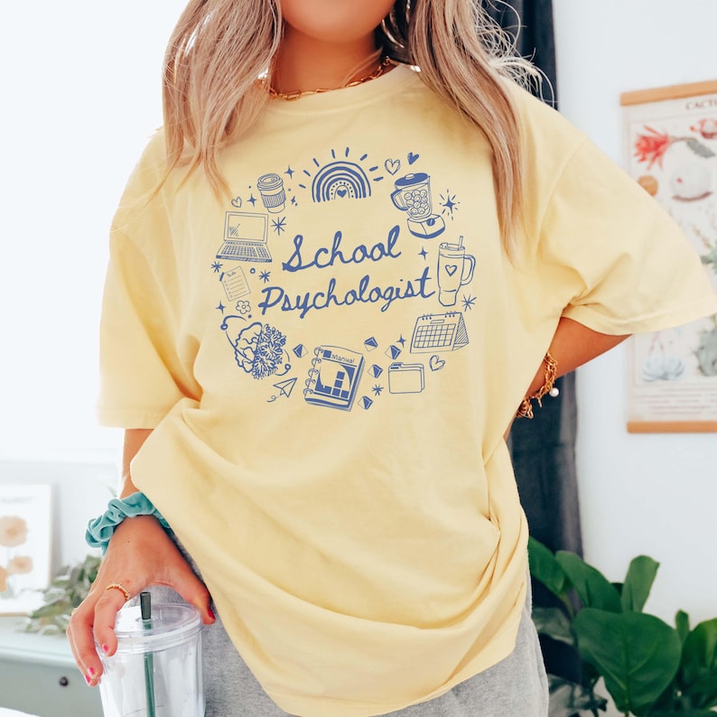 May include: A light yellow t-shirt with a blue graphic that says "School Psychologist." The graphic includes a laptop, coffee cup, rainbow, and other school-related icons. The shirt is a comfortable, oversized fit.