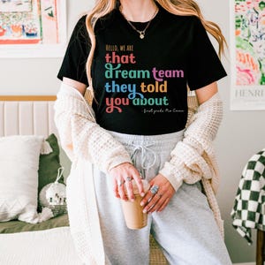 May include: Black t-shirt with the text "HELLO, WE ARE that dream team they told you about - first grade Mo Emma" in colorful lettering. The shirt is paired with gray sweatpants and a cream-colored cardigan.