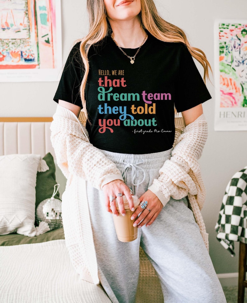 May include: Black t-shirt with the text "HELLO, WE ARE that dream team they told you about - first grade Mo Emma" in colorful lettering. The shirt is paired with gray sweatpants and a cream-colored cardigan.