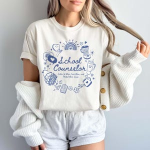 May include: A white t-shirt featuring a blue graphic design that says "School Counselor" and includes various illustrations such as hearts, a rainbow, a watering can, and a blender. The text below the title reads "Listen to them, Love them, and Watch them Grow."