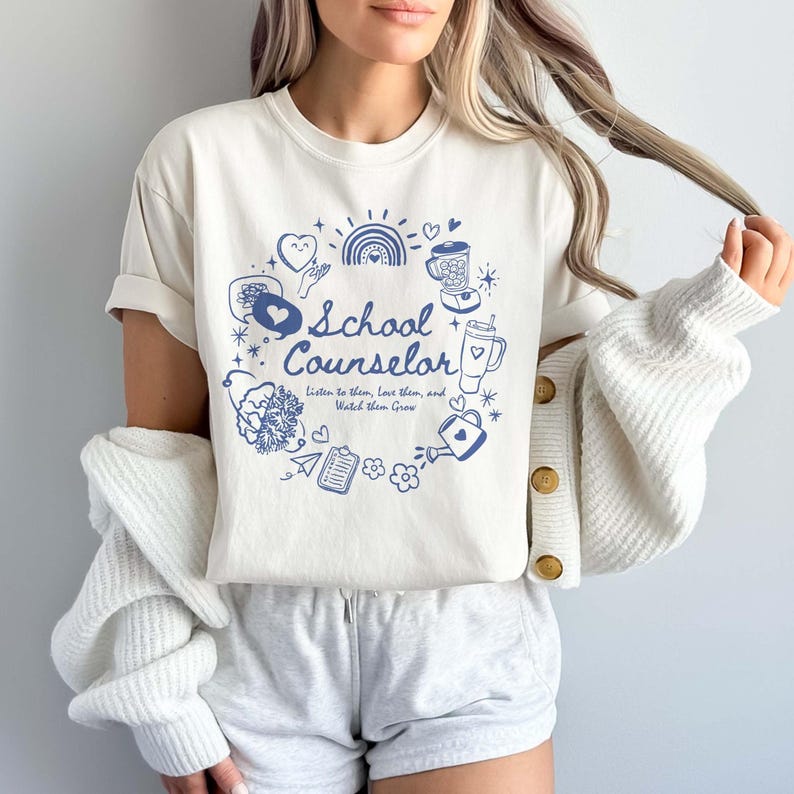May include: A white t-shirt featuring a blue graphic design that says "School Counselor" and includes various illustrations such as hearts, a rainbow, a watering can, and a blender. The text below the title reads "Listen to them, Love them, and Watch them Grow."