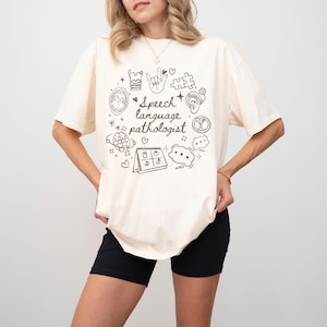 K&ouml;nnte beinhalten: Eine Frau tr&auml;gt ein hellbeiges T-Shirt mit einem braunen Grafikdesign, auf dem "Speech language pathologist" steht und verschiedene Symbole im Zusammenhang mit Logop&auml;die enthalten sind. Sie tr&auml;gt eine schwarze Shorts.