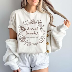 May include: A white t-shirt with a brown graphic design that says "Social Worker" surrounded by various illustrations. The text below the graphic says "Empathy, Cultural Awareness, Compassion, Advocacy, Communication, Support".