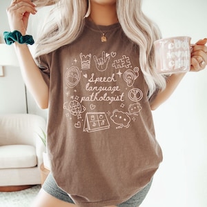 K&ouml;nnte beinhalten: Ein braunes T-Shirt mit einem wei&szlig;en Grafikdesign, das den Text "Speech Language Pathologist" zeigt, umgeben von verschiedenen Symbolen, die mit Logop&auml;die zu tun haben, wie z. B. ein Gehirn, ein Kalender und eine Sprechblase.