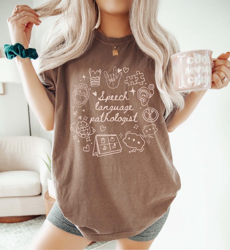 K&ouml;nnte beinhalten: Ein braunes T-Shirt mit einem wei&szlig;en Grafikdesign, das den Text "Speech Language Pathologist" zeigt, umgeben von verschiedenen Symbolen, die mit Logop&auml;die zu tun haben, wie z. B. ein Gehirn, ein Kalender und eine Sprechblase.