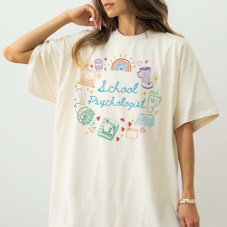 May include: Cream-colored t-shirt with a circular design featuring the words "School Psychologist" in blue script. The design includes colorful illustrations of a rainbow, coffee cup, laptop, blender, and other related icons.