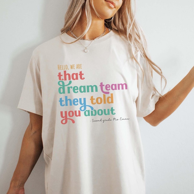 May include: Cream-colored t-shirt with the text "HELLO, WE ARE that dream team they told you about." The text is in various colors, including red, green, purple, and orange. "Second grade Ms. Emma" is written below the main text.