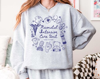 May include: Gray sweatshirt with a blue design featuring the text "Neonatal Intensive Care Unit" and various illustrations related to newborn care, including a stethoscope, a baby bottle, a pacifier, a baby onesie, a heart rate monitor, and a teddy bear.