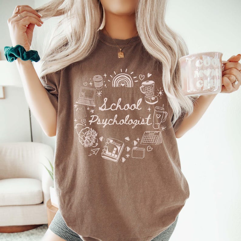 May include: A brown t-shirt with the words "School Psychologist" in a white circular design. The design includes a rainbow, coffee cup, laptop, and other small illustrations. A pink mug with the words "Cool Moms Club" is held in the hand.