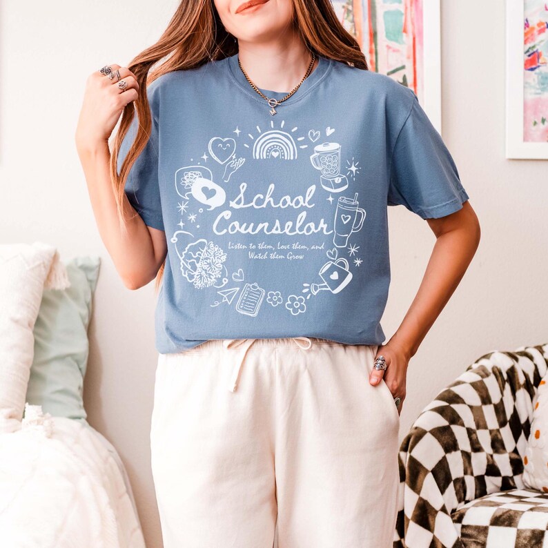May include: A light blue t-shirt with a white graphic design that says "School Counselor" surrounded by various illustrations. The text below the graphic says "Listen to them, love them, and watch them grow."