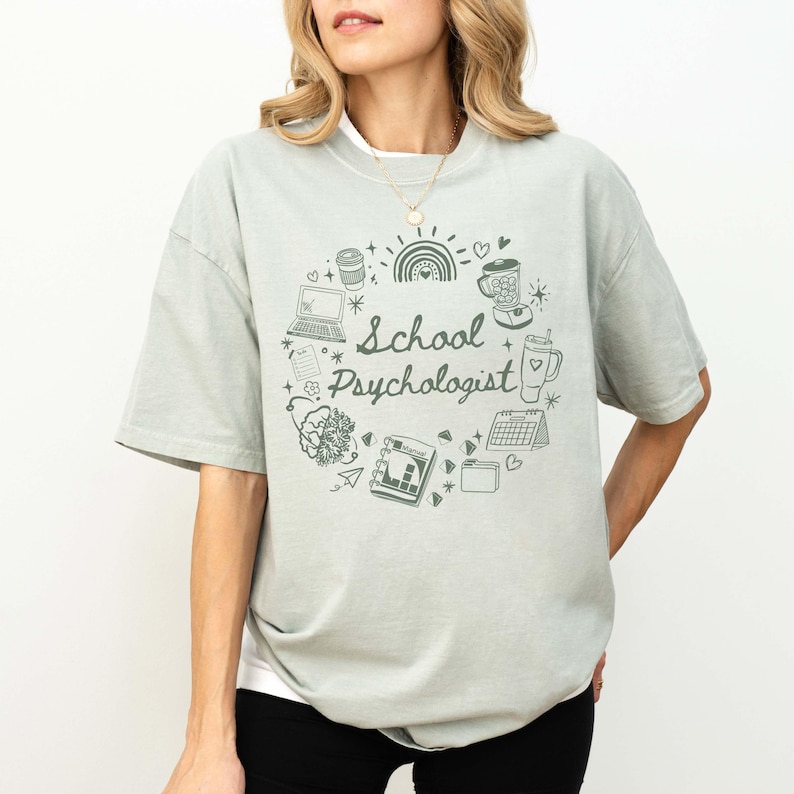 May include: A light green t-shirt with the words "School Psychologist" in a circular design. The design includes a laptop, coffee cup, rainbow, and other small illustrations. The shirt is a relaxed fit.