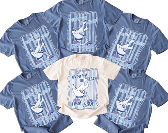May include: Several blue and cream-coloured t-shirts. Each shirt displays a graphic of a goose holding a pencil, with the text "On My Way To Teach" and "First Grade" within a blue frame. These are casual wear items.