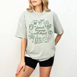 K&ouml;nnte beinhalten: Ein hellgr&uuml;nes T-Shirt mit einem grafischen Design von Logop&auml;die-Symbolen und dem Text "Speech language pathologist".