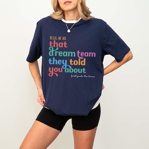 May include: Navy blue t-shirt with the text "HELLO, WE ARE that dream team they told you about" in colorful lettering. The shirt also includes the text "first grade Mo Emma". The shirt is a casual style.