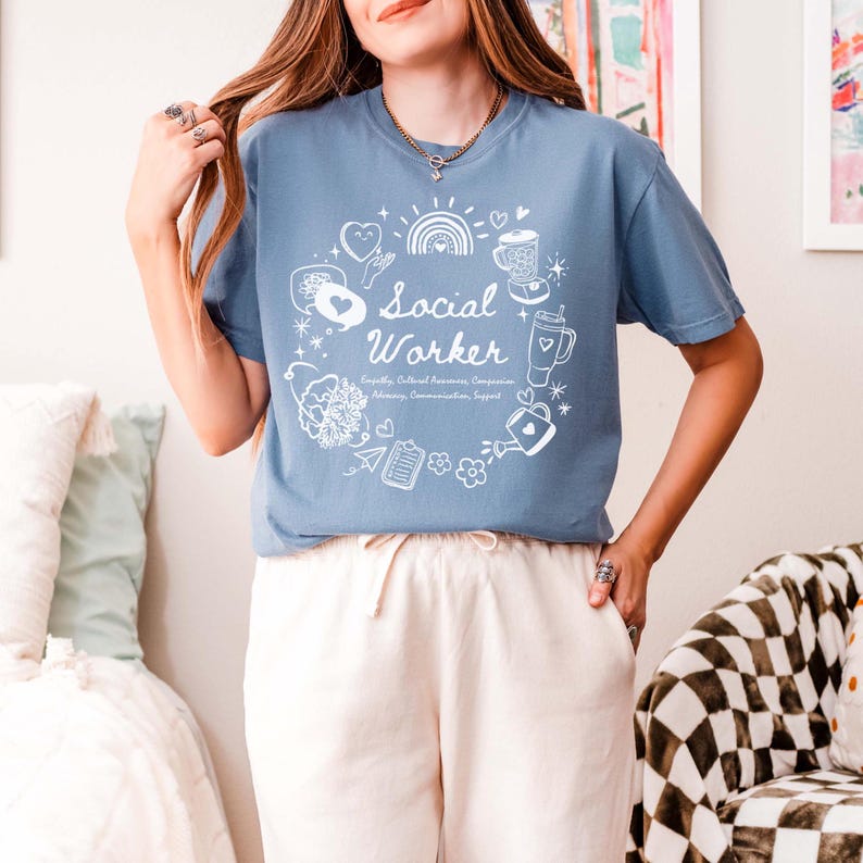 May include: A light blue t-shirt with a white graphic design. The graphic features a circle with the words "Social Worker" and various illustrations of hearts, a rainbow, a coffee cup, a watering can, and other objects. The text below the circle reads "Empathy, Cultural Awareness, Compassion, Advocacy, Communication, Support."