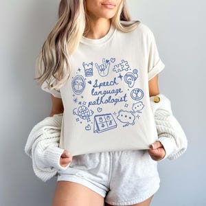 K&ouml;nnte beinhalten: Ein wei&szlig;es T-Shirt mit einem blauen Design, das verschiedene Symbole im Zusammenhang mit Logop&auml;die zeigt, darunter ein Gehirn, ein Mund, eine Hand, ein Puzzle-Teil und einen Kalender. Der Text "Speech language pathologist" ist in Blau auf dem Shirt geschrieben.