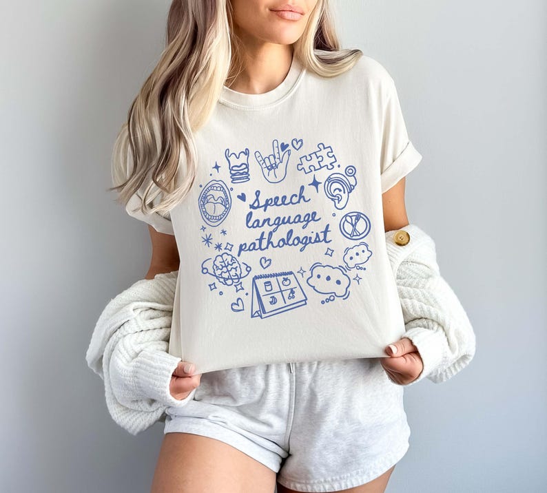 K&ouml;nnte beinhalten: Ein wei&szlig;es T-Shirt mit einem blauen Design, das verschiedene Symbole im Zusammenhang mit Logop&auml;die zeigt, darunter ein Gehirn, ein Mund, eine Hand, ein Puzzle-Teil und einen Kalender. Der Text "Speech language pathologist" ist in Blau auf dem Shirt geschrieben.