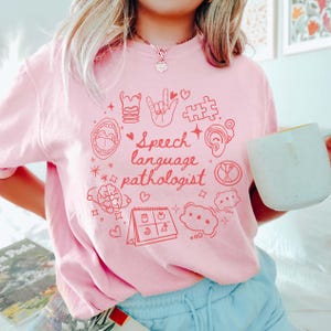 K&ouml;nnte beinhalten: Pinkes T-Shirt mit einem roten Grafikdesign mit dem Text "Speech language pathologist" und verschiedenen Symbolen im Zusammenhang mit Logop&auml;die, darunter ein Gehirn, ein Kalender, ein Puzzle-Teil und eine Hand, die das Zeichen f&uuml;r "Ich liebe dich" macht.