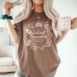 May include: A brown t-shirt with a white graphic design that says "School Counselor" with a variety of illustrations surrounding the text. The illustrations include hearts, a rainbow, a watering can, a flower, a pencil, a clipboard, a paper aeroplane, and a coffee cup.
