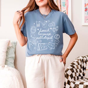 K&ouml;nnte beinhalten: Ein hellblaues T-Shirt mit einem wei&szlig;en Design aus Logop&auml;die-Symbolen und dem Text "Speech language pathologist".