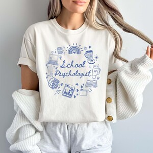 May include: Cream-colored t-shirt featuring a blue circular design with the words "School Psychologist." The design includes illustrations of a laptop, coffee cup, rainbow, and other related icons. The shirt is part of a casual wear collection.