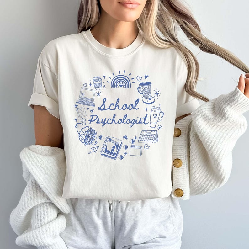 May include: Cream-colored t-shirt featuring a blue circular design with the words "School Psychologist." The design includes illustrations of a laptop, coffee cup, rainbow, and other related icons. The shirt is part of a casual wear collection.