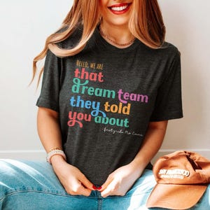May include: Dark gray t-shirt with the text "HELLO, WE ARE that dream team they told you about" in colorful, playful lettering. The shirt is a crew neck and is paired with blue jeans and a brown baseball cap.