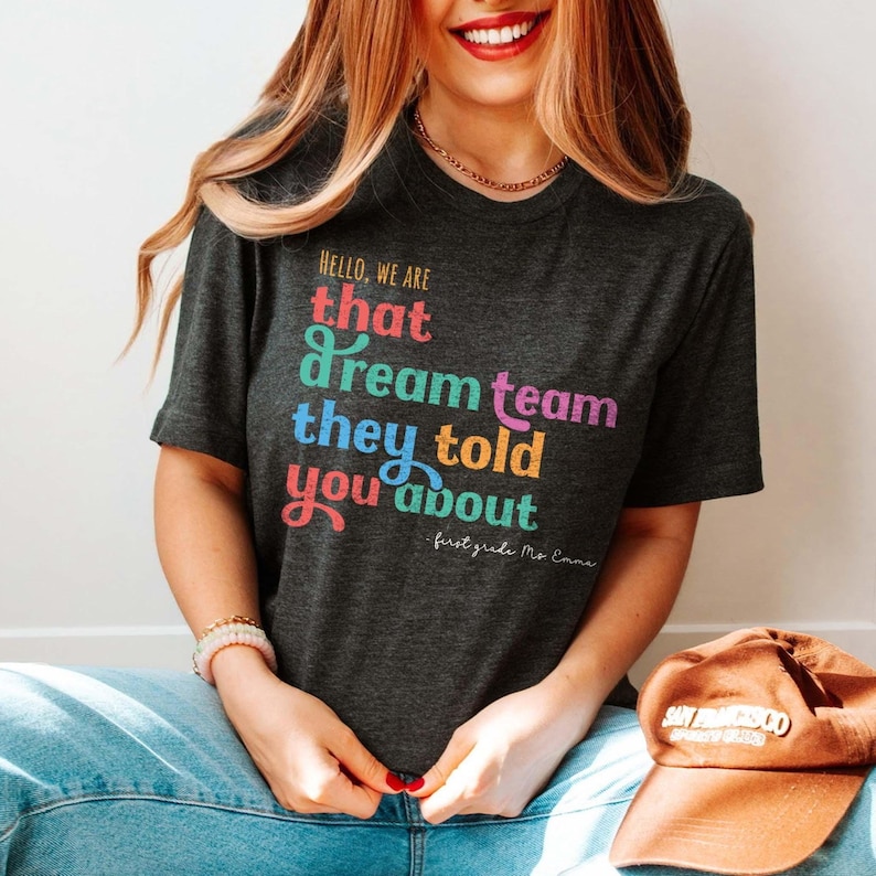 May include: Dark gray t-shirt with the text "HELLO, WE ARE that dream team they told you about" in colorful, playful lettering. The shirt is a crew neck and is paired with blue jeans and a brown baseball cap.