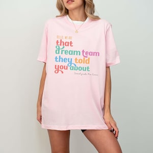May include: A light pink t-shirt with the text "HELLO, WE ARE that dream team they told you about" in various colors. The text "Second grade Ms. Emma" is below the main text. The shirt has short sleeves.