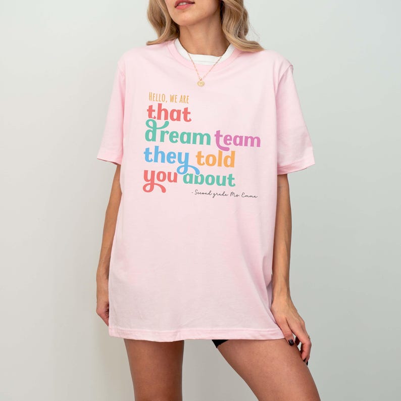 May include: A light pink t-shirt with the text "HELLO, WE ARE that dream team they told you about" in various colors. The text "Second grade Ms. Emma" is below the main text. The shirt has short sleeves.