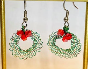 May include: Handmade earrings with green, open-weave wreaths and red bead accents. The earrings hang from silver-coloured hooks, creating a festive holiday accessory. The wreaths are approximately 3.8 cm in diameter.