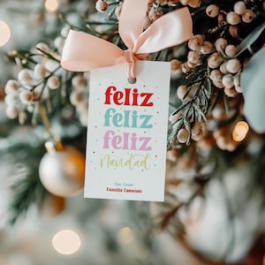 Puede incluir: Una etiqueta de regalo blanca con las palabras "Feliz Feliz Feliz Navidad" en rojo, azul y rosa, atada con una cinta rosa. La etiqueta está colgada en un árbol de Navidad con bayas blancas y una bola dorada.