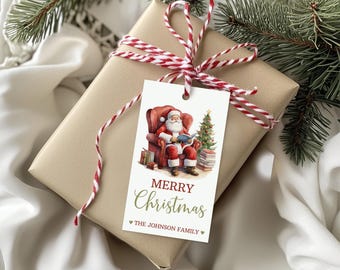 May include: A white gift tag with a red and white striped twine bow. The tag features a watercolour illustration of Santa Claus sitting in a red armchair reading a book. A Christmas tree and a stack of books are beside him. The text on the tag reads "Merry Christmas" in green script and "The Johnson Family" in a smaller font.