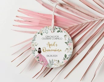 May include: A round tag with the text "Gracias por celebrar conmigo" and "April's Quinceañera" in gold script. The tag features a watercolor illustration of a girl in a pink dress and is decorated with floral elements. The date 06.28.202X is also included.