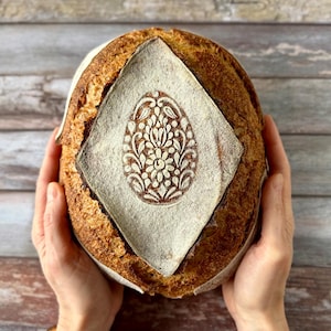 May include: A round loaf of artisan bread with a decorative egg design on top. The bread is held by two hands, showcasing the intricate details of the baked goods. The crust is golden brown, and the design is a lighter colour.