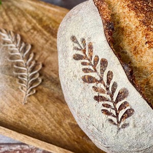 May include: A freshly baked loaf of bread with a leaf pattern dusted in flour. The bread has a light crust with a dark, textured top. Two leaf designs are also dusted in flour on a wooden tray.