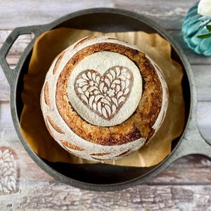 May include: A round loaf of artisan bread with a heart-shaped design, baked in a cast iron skillet. The bread is golden brown with a decorative flour pattern. A heart-shaped stencil is visible on the wooden surface.