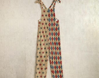 May include: A tan overall jumpsuit featuring red, blue, and tan star and diamond patterns. The straps are tied at the shoulders with matching fabric. The jumpsuit is hanging on a hanger against a neutral background.