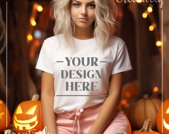 Könnte beinhalten: Ein weißes T-Shirt mit dem Text "YOUR DESIGN HERE" auf der Vorderseite. Das T-Shirt wird von einer Person mit blonden Haaren getragen, die vor einem Hintergrund aus Kürbissen und Lichtern sitzt.