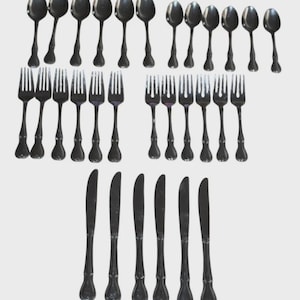 Puede incluir: Una colección de cubiertos negros, que incluye cucharas, tenedores y cuchillos, dispuestos sobre un fondo blanco. Las cucharas tienen cuencos redondeados, los tenedores tienen cuatro púas y los cuchillos tienen una hoja curva. Los cubiertos parecen ser de metal.