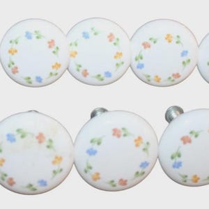 May include: A set of ten round white ceramic cabinet knobs, each featuring a hand-painted circular wreath of small orange, blue, and green floral motifs. Each knob includes a silver-toned metal screw attachment extending from the back side.