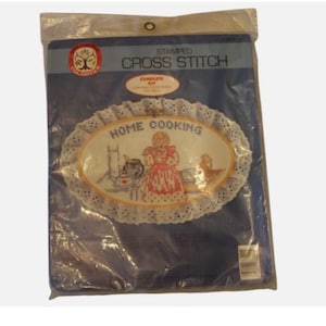 May include: A blue package containing a stamped cross stitch kit. The kit features an oval design with the words "HOME COOKING" and a depiction of a woman in a red dress. The design is surrounded by a white lace trim.
