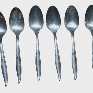 May include: Six silver spoons displayed in a line. Each spoon features an oval bowl and a thin handle with a floral pattern. The spoons are made of a shiny, reflective metal.