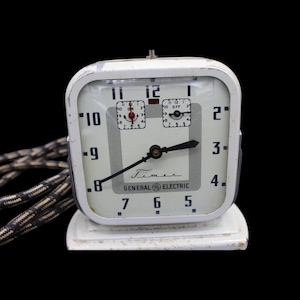Puede incluir: Un reloj temporizador General Electric blanco vintage con una esfera cuadrada. El reloj tiene manecillas negras, un cable en blanco y negro y la palabra "Timer" está impresa en la esfera. El reloj está ajustado a las 6:30.