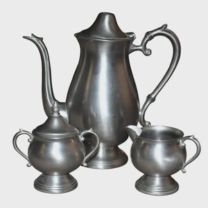 Puede incluir: Un juego de té de peltre vintage, que incluye una cafetera alta con un pico curvo, un azucarero con tapa y una pequeña jarra para crema. El juego tiene un diseño clásico con asas ornamentadas y un acabado pulido.