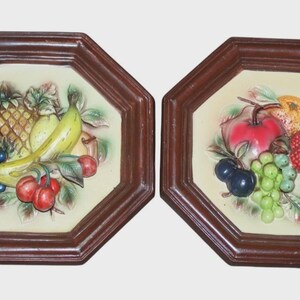 Puede incluir: Dos placas de pared octogonales con marcos de madera marrón. Cada placa presenta un relieve de frutas coloridas, incluyendo uvas, plátanos, manzanas y bayas. El fondo es de color crema.