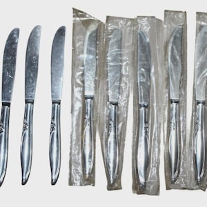 May include: A set of stainless steel knives, some individually wrapped in clear plastic. The knives have a decorative floral design on the handles. The blades are curved and appear sharp.