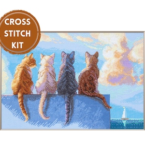 May include: A cross stitch kit featuring a needlepoint design of four cats sitting on a blue wall, facing a sea with a small sailboat under a cloudy sky. The cats are orange, white, dark grey, and brown. A brown circular badge says CROSS STITCH KIT.
